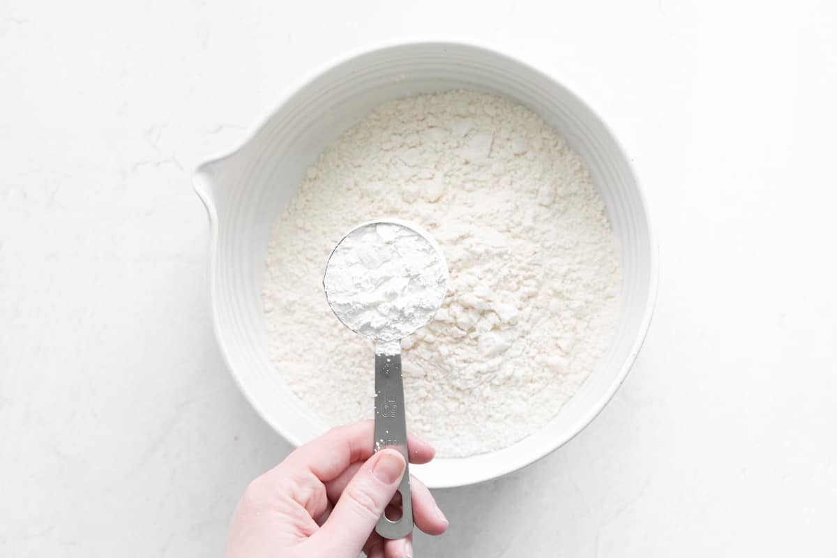 hand holding a measuring cup of cornstarch above a bowl of flour
