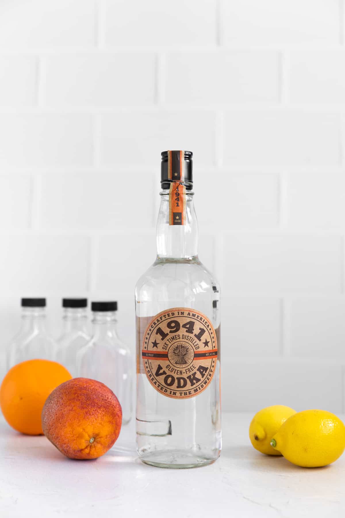 a bottle of vodka at the center, surrounded by various citrus fruits, against a white tiled background