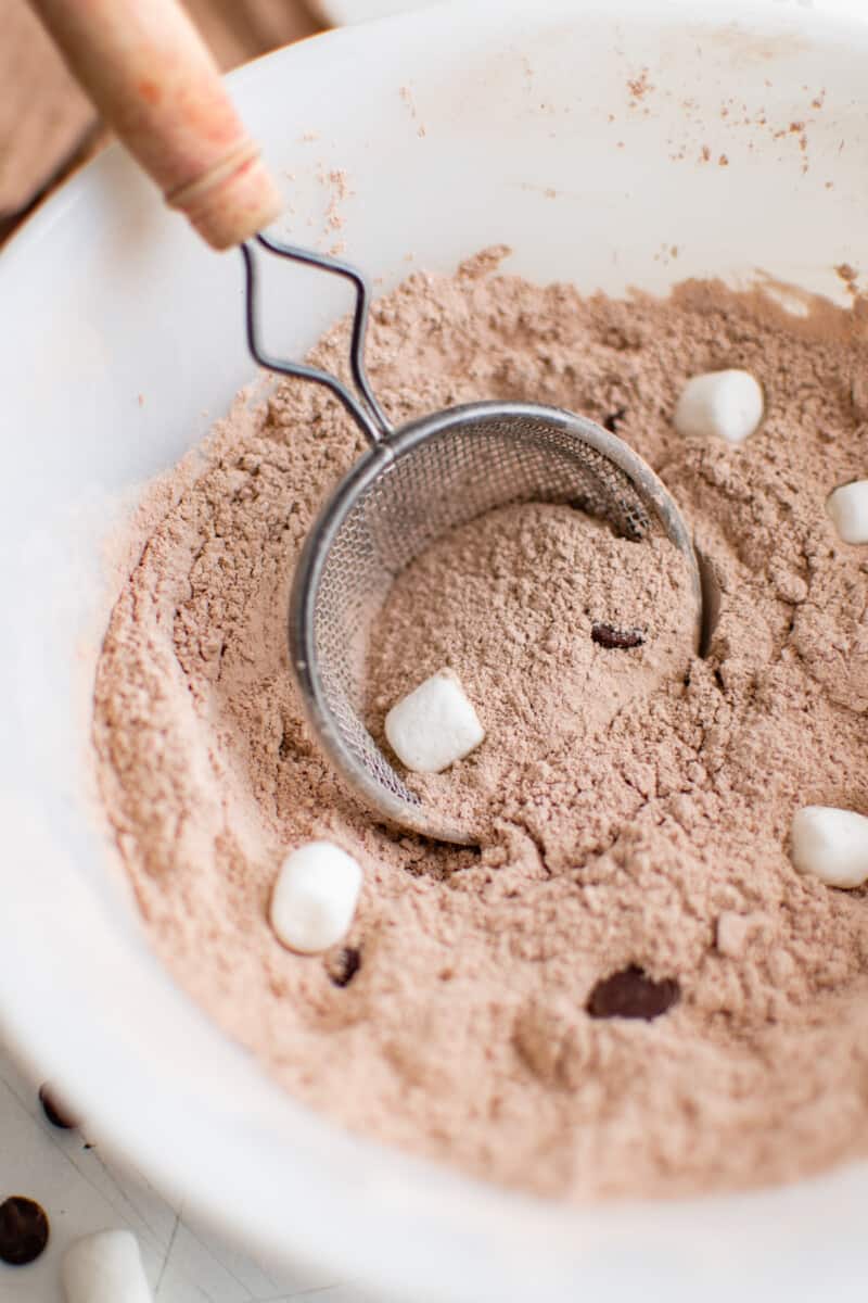 a sifter scooping homemade hot chocolate mix from a white bowl.