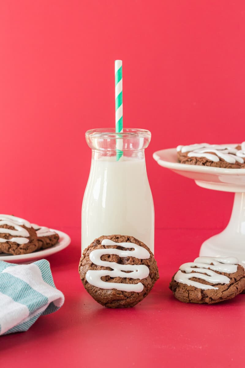 a hot cocoa cookie leaning against a glass of milk with a green and white straw.