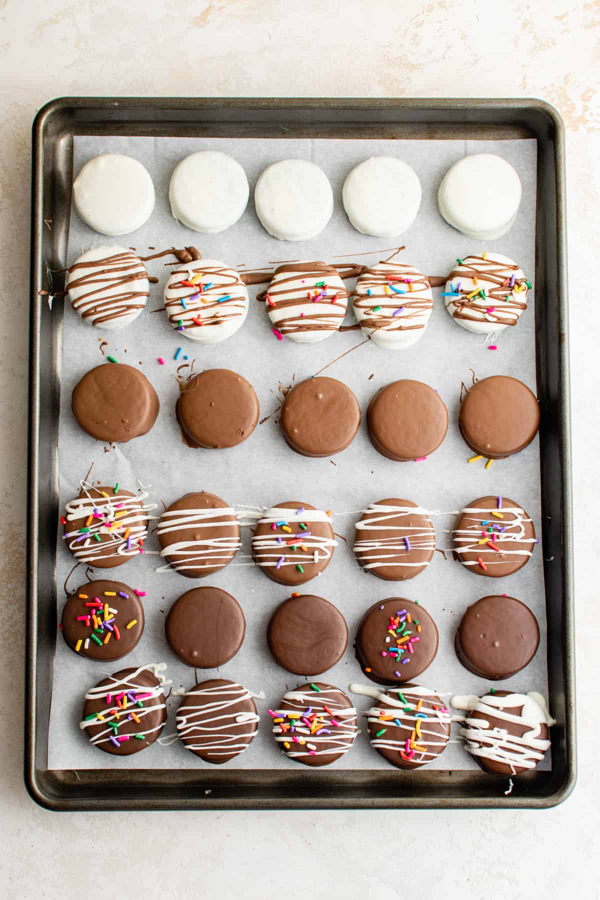 chocolate covered Oreos lined up on a baking tray; some are white chocolate, some are dark chocolate, some are milk chocolate; some are plain, some are drizzled with more chocolate, some have sprinkles