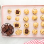 balls of cookie dough arranged on a baking tray, cubes of brownies being placed at the center of some of the cookies