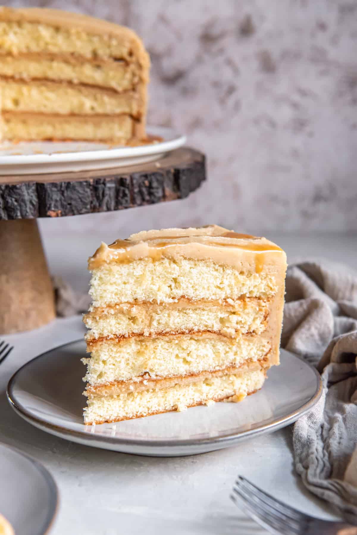 a slice of layered caramel cake on a plate, the rest of the cake is on a cake stand in the background
