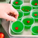 a hand grabbing 1 of 8 grinch jello shots from a rimmed baking sheet.