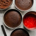 overhead view of 3 layers of chocolate strawberry cake next to filling and frosting in stainless bowls.