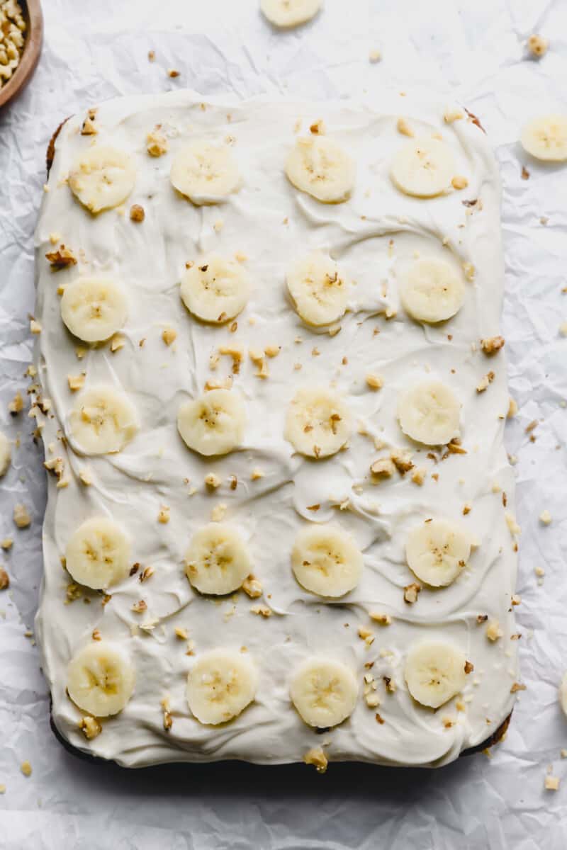 overhead view of banana cake on a white table with banana slices and crushed nuts on top.
