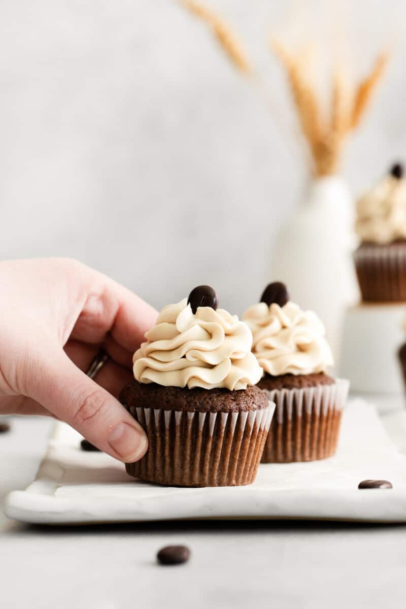 a hand grabbing 1 of 2 kahlua cupcakes from a rectangular white plate.