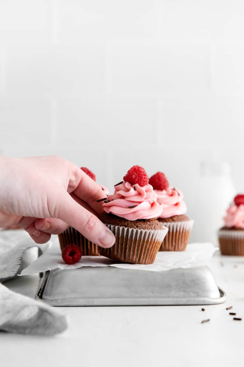 a hand grabbing 1 of 3 chocolate raspberry cupcakes from an overturned square cake tin.