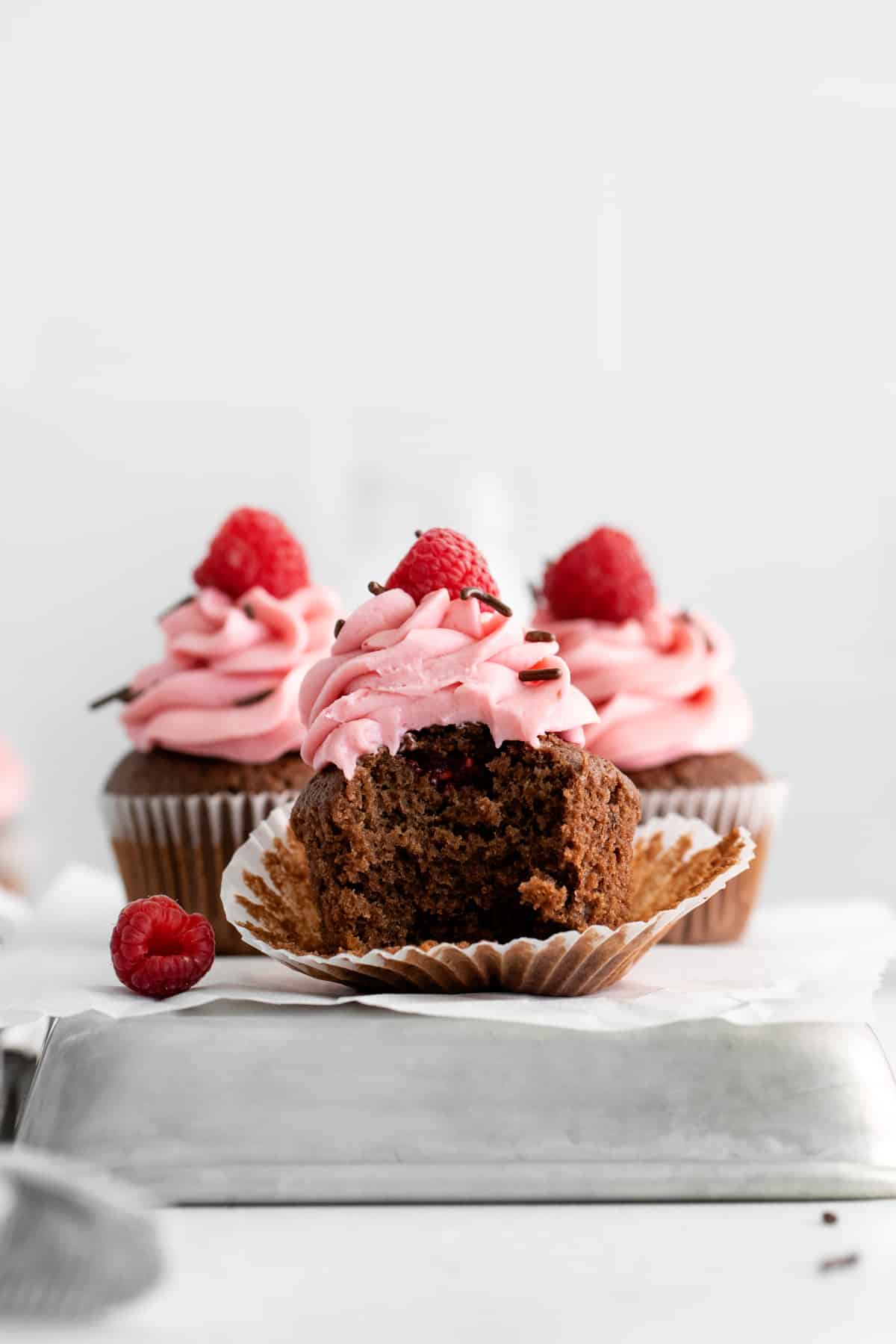 a bitten and unwrapped chocolate raspberry cupcake on an overturned square cake pan with 2 cupcakes behind it.