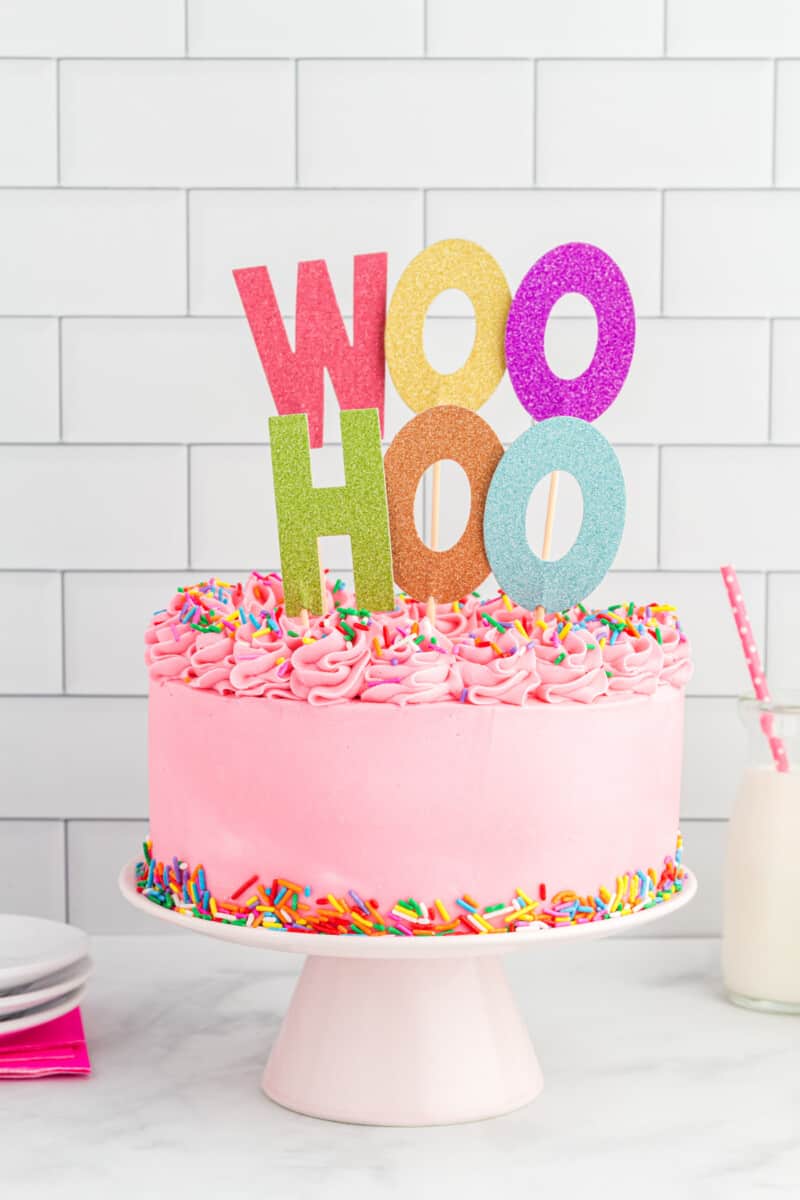a pink frosted cake on a white cake stand with a topper that reads woo hoo in rainbow letters.