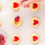 a hand using a spoon to add jam to heart thumbprint cookies.