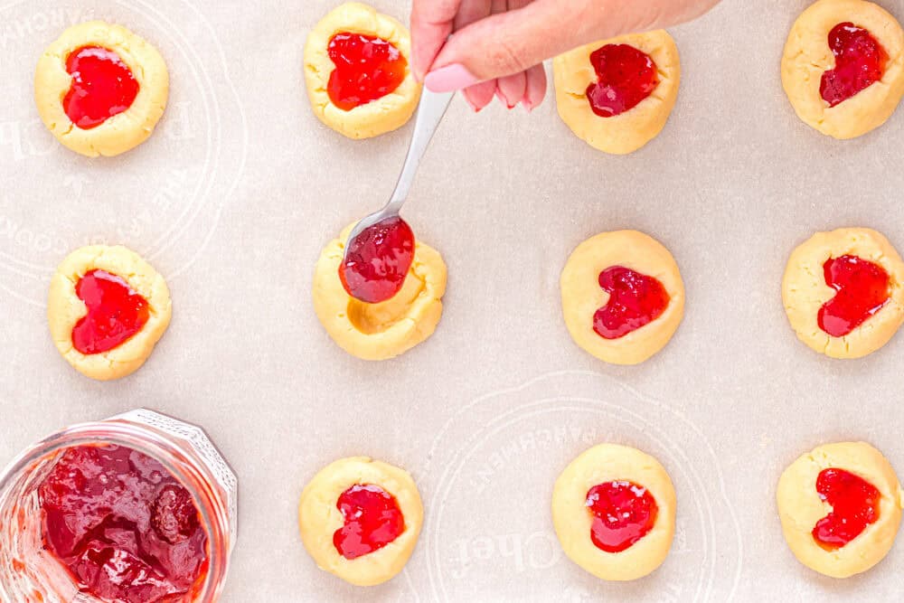 a hand using a spoon to add jam to heart thumbprint cookies.