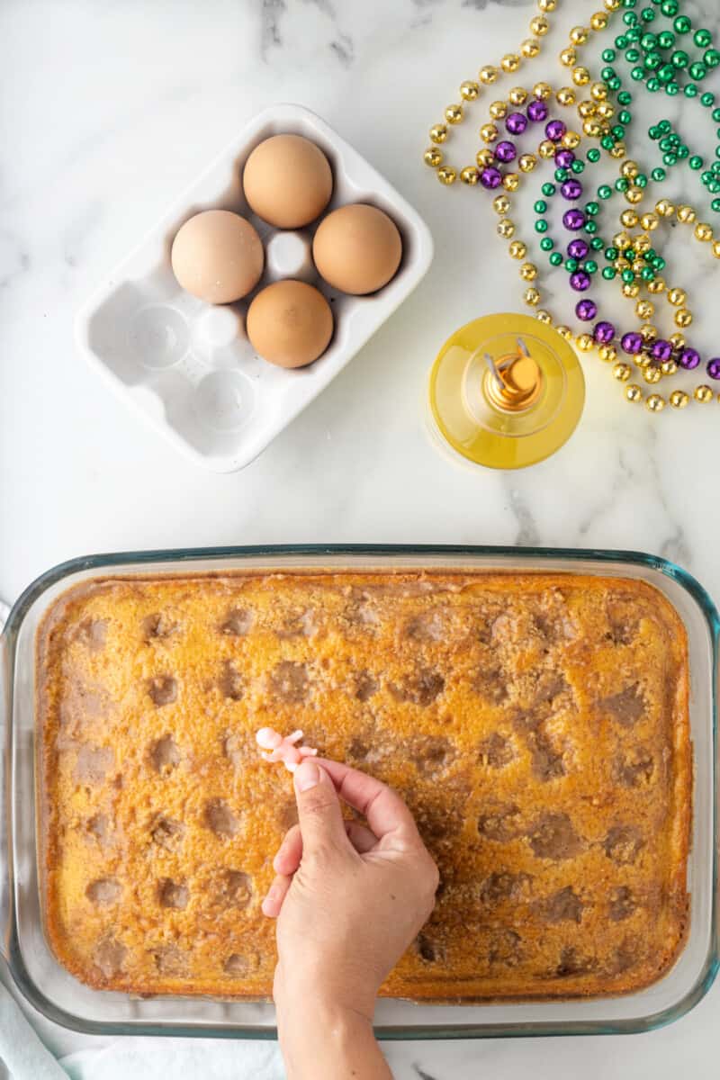 a hand holding a plastic baby over a set king cake poke cake in a rectangular baking pan.