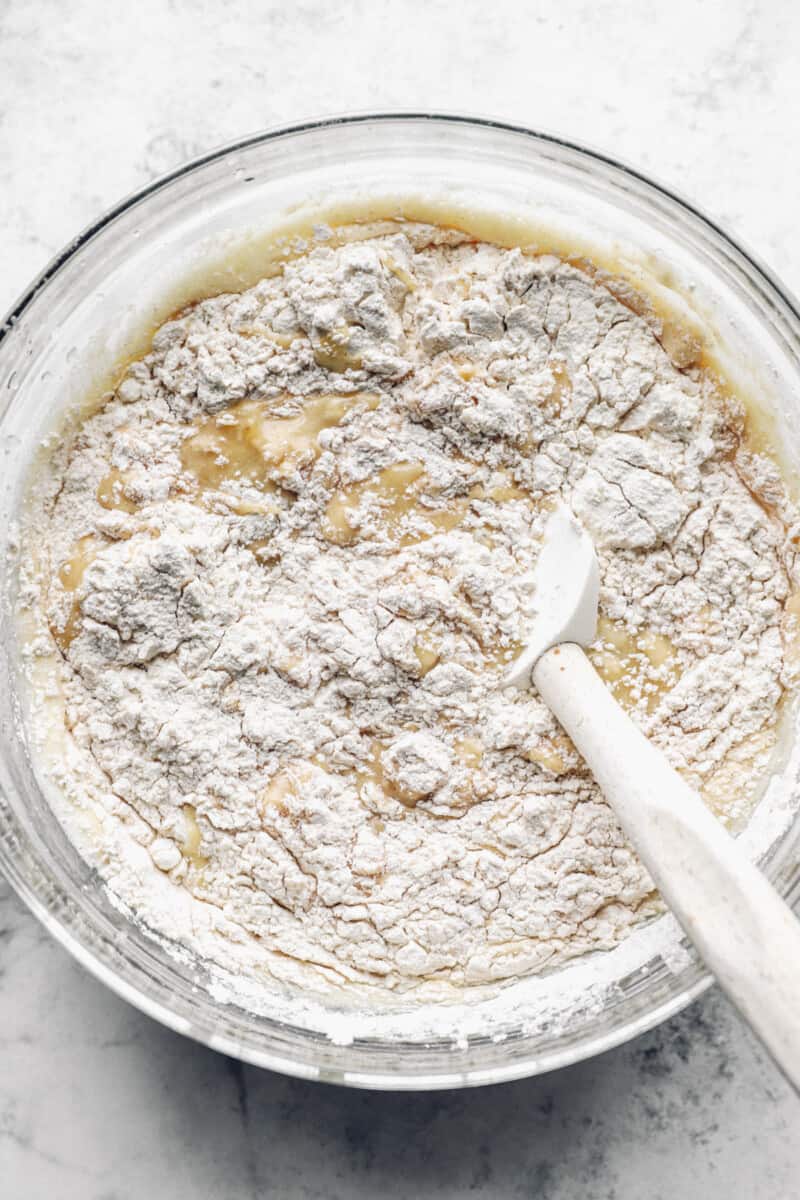 dry ingredients being folded into wet ingredients for banana cake in a glass bowl with a rubber spatula.