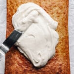 an offset spatula spreading cream cheese frosting over a baked banana cake.