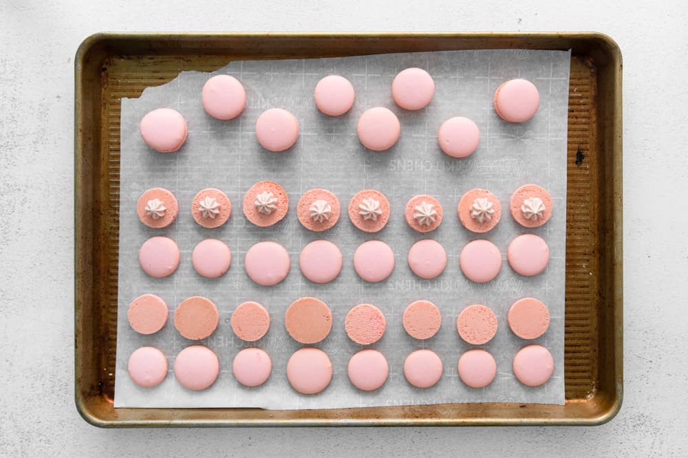 baked raspberry macaron shells on a baking sheet, half flipped over to expose the underside and dolloped with buttercream.