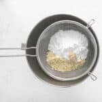 dry ingredients for macarons in a fine-mesh sieve set over a stainless mixing bowl.