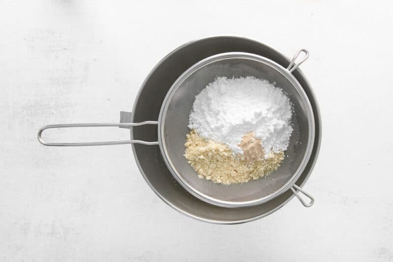 dry ingredients for macarons in a fine-mesh sieve set over a stainless mixing bowl.
