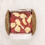 overhead view of cheesecake dolloped on red velvet brownie batter in a square baking pan.