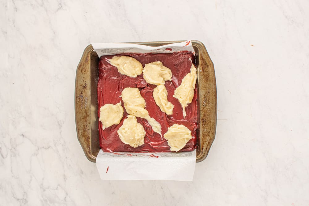 overhead view of cheesecake dolloped on red velvet brownie batter in a square baking pan.