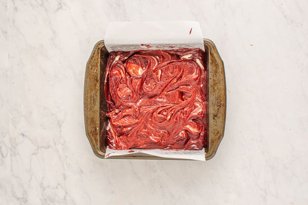 overhead view of cheesecake swirled into red velvet brownie batter in a square baking pan.