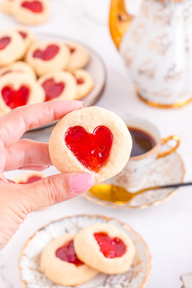 a hand holding a strawberry heart thumbprint cookie between the thumb and pointer finger.