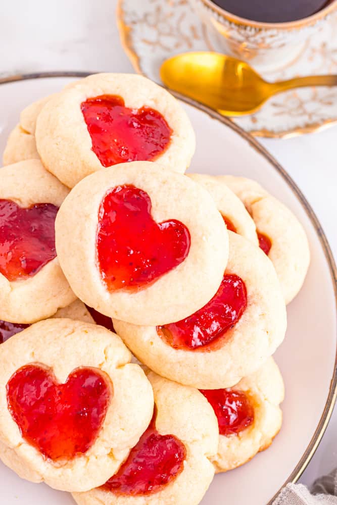 partial overhead view of heart thumbprint cookies filled with strawberry jam on a white plate.