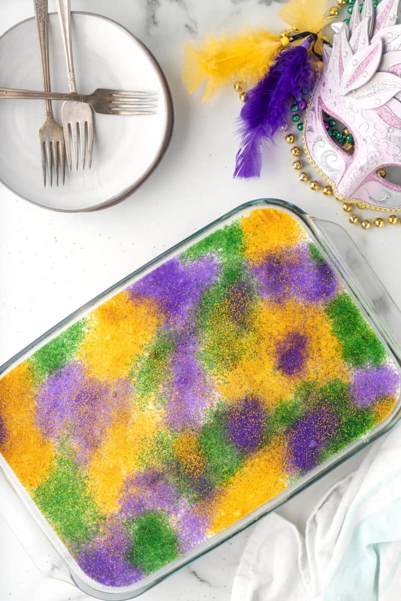 overhead view of king cake poke cake next to a plate with forks and a mask.