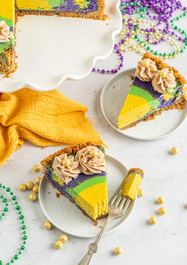 slices of king cake cheesecake on white plates, one missing a bite with a fork.