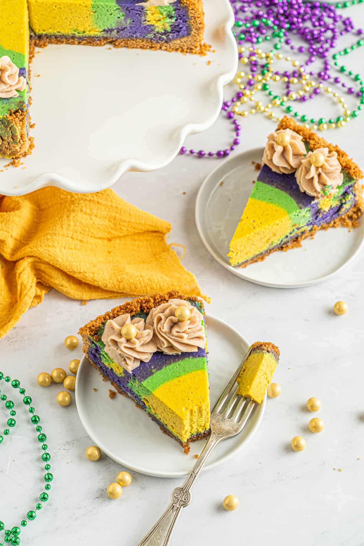 slices of king cake cheesecake on white plates, one missing a bite with a fork.