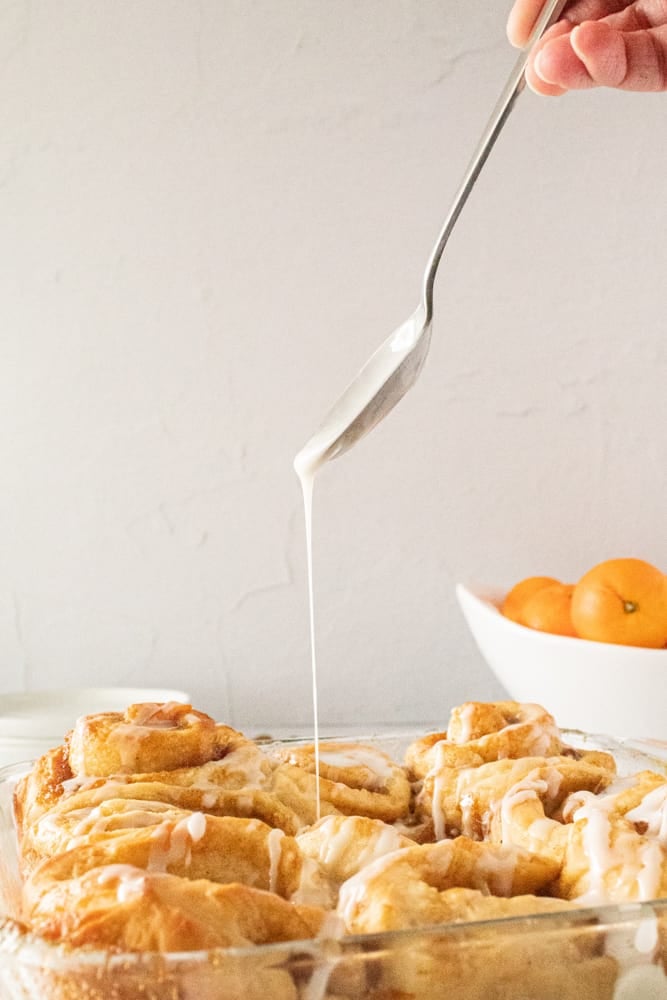 spoon drizzling icing over freshly baked orange rolls