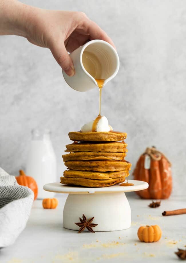 a person pouring syrup on a stack of pumpkin pancakes.