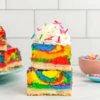 two stacked rainbow cheesecake bars topped with whipped cream and rainbow sprinkles.