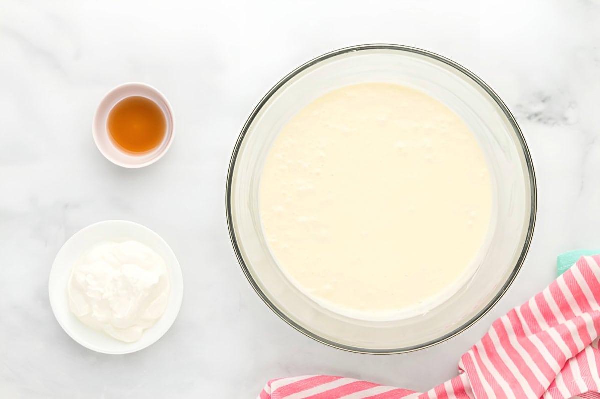 cheesecake batter in a bowl with bowls of sour cream and vanilla extract next to it.