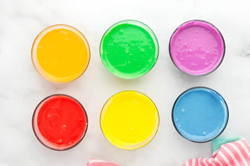 six glass bowls of cheesecake batter, each colored a different color of the rainbow.