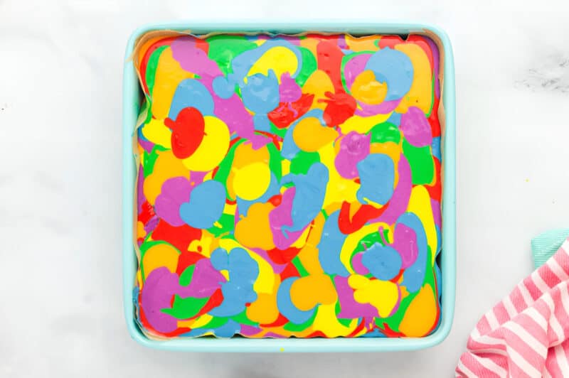 swirled rainbow cheesecake batter in a lined square baking pan.