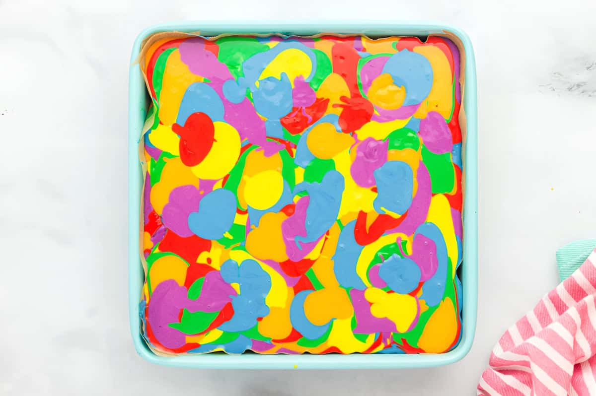 swirled rainbow cheesecake batter in a lined square baking pan.