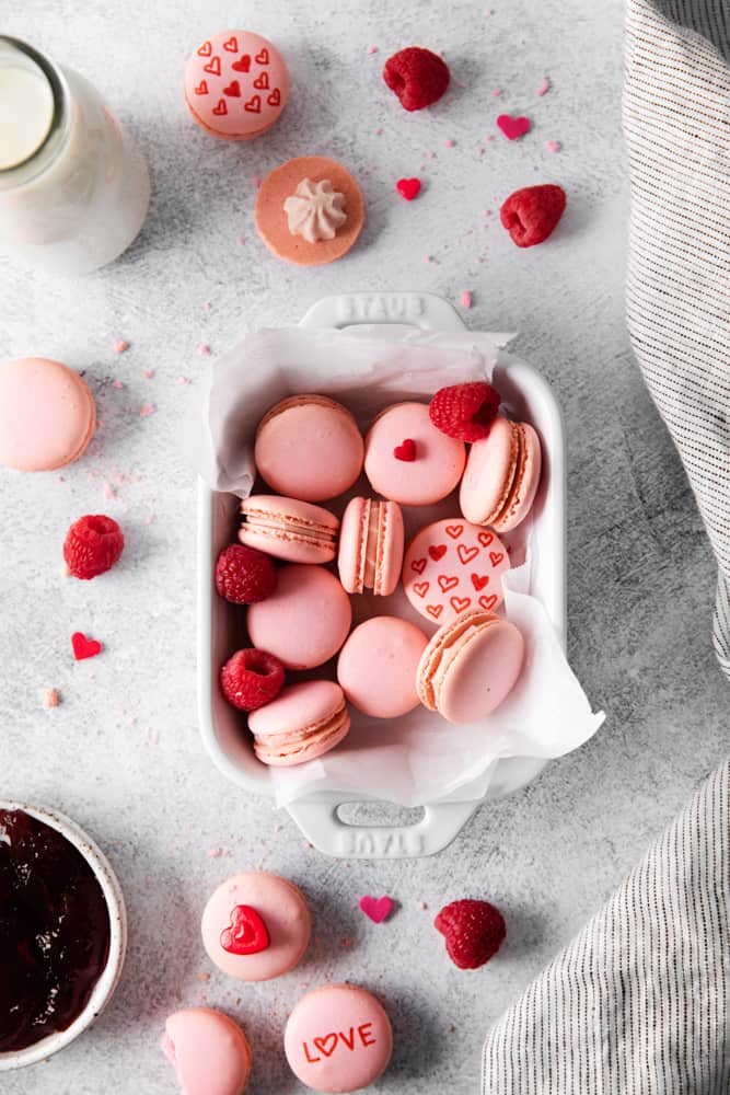 overhead view of raspberry macarons in a parchment-lined white rectangular serving dish with hearts and writing on them.