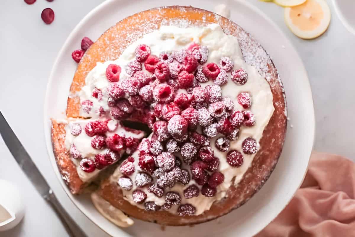 overhead view of raspberry olive oil cake topped with whipped cream and fresh raspberries on a cake stand.