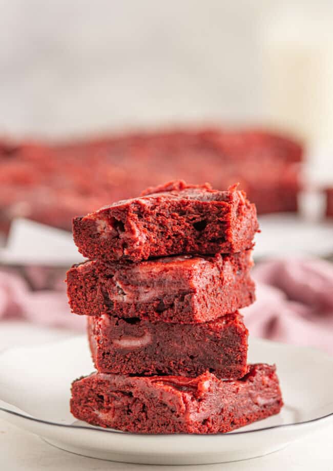 close up side view of a partially eaten red velvet cheesecake brownie on top of a stack of 3 red velvet cheesecake brownies on a white plate.