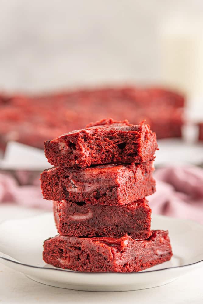 close up side view of a partially eaten red velvet cheesecake brownie on top of a stack of 3 red velvet cheesecake brownies on a white plate.