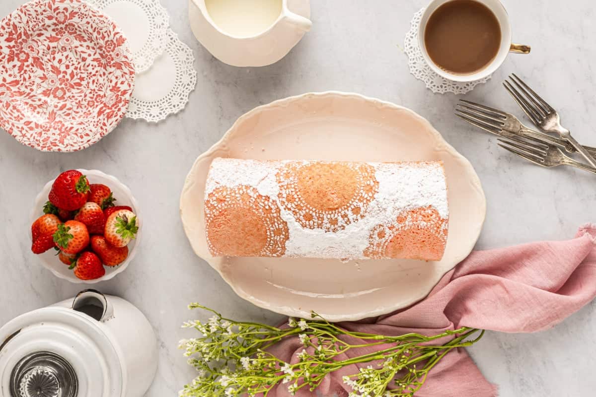 overhead view of strawberry roll cake on a pink platter with a powdered sugar doily design on top.