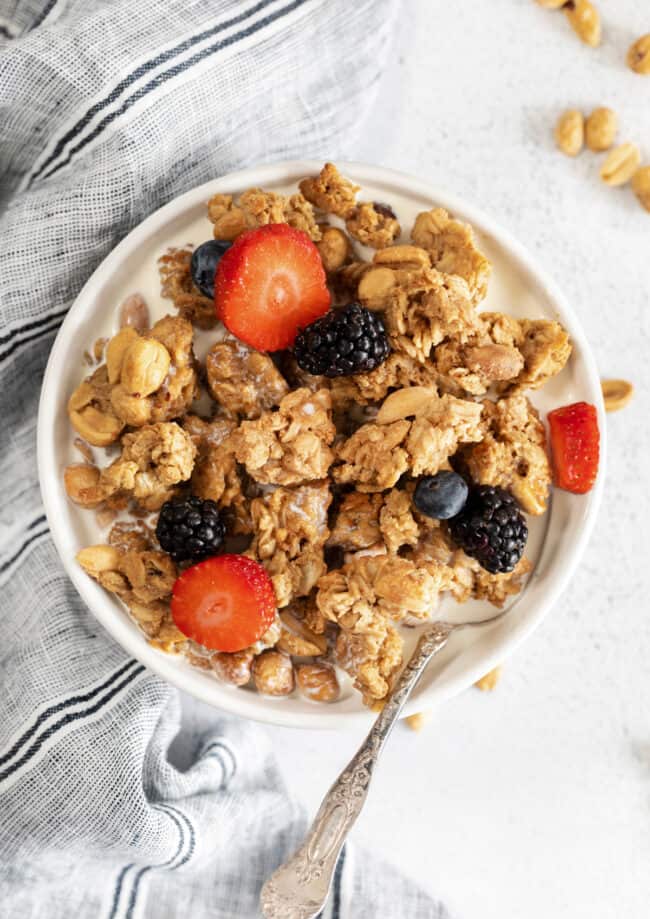 overhead view of a serving of peanut butter granola in a white bowl with a spoon and berries on top.