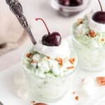 a cup of watergate salad with a spoon, topped with crushed nuts, whipped cream, and a cherry.