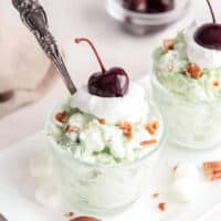 a cup of watergate salad with a spoon, topped with crushed nuts, whipped cream, and a cherry.