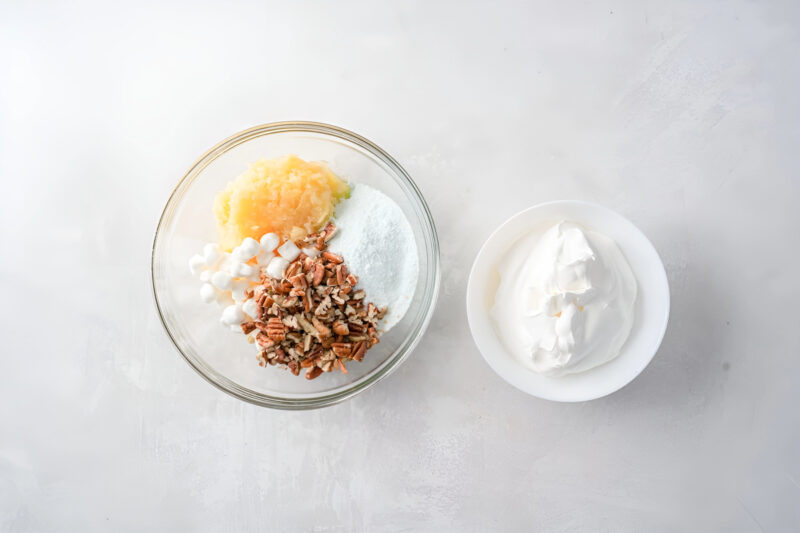 crushed pineapple, pudding mix, chopped nuts, and mini marshmallows in a bowl next to a bowl of whipped topping.