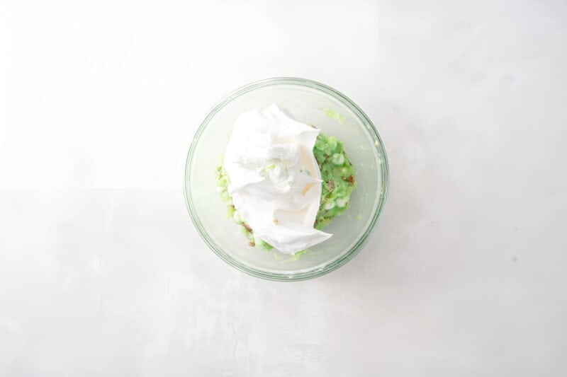 adding whipped topping to pistachio fruit and nut salad in a glass bowl.
