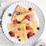 four folded sweet crepes topped with powdered sugar and fresh berries on a plate.