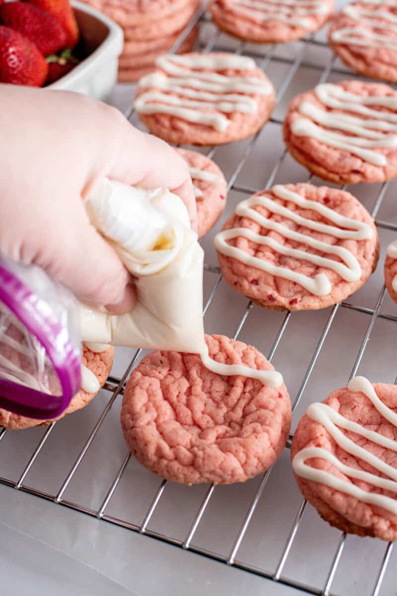 a hand piping cream cheese glaze over strawberry cookies on a wire raack.