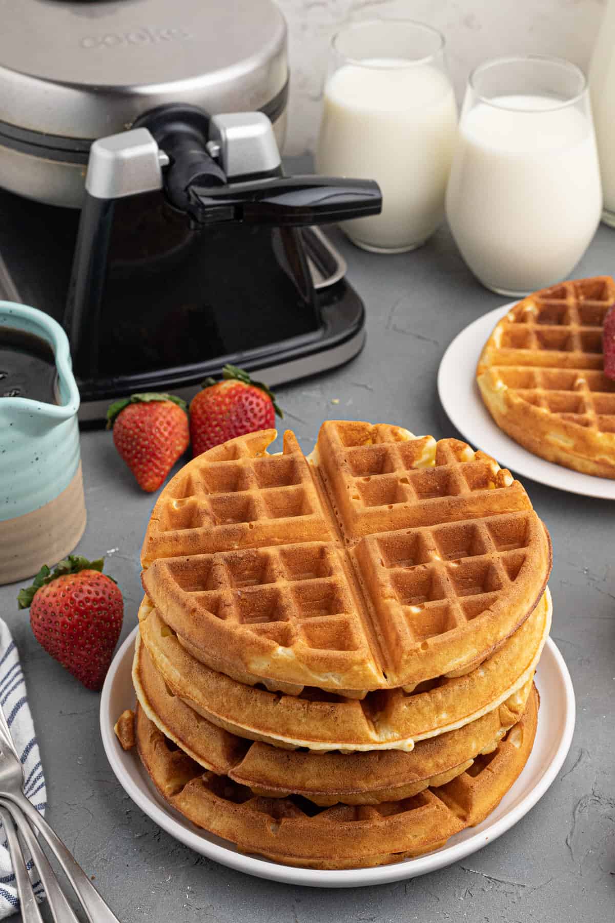 a stack of waffles next to a waffle maker, with glasses of milk in the background